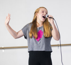 Teen girl belting into a microphone on stage.